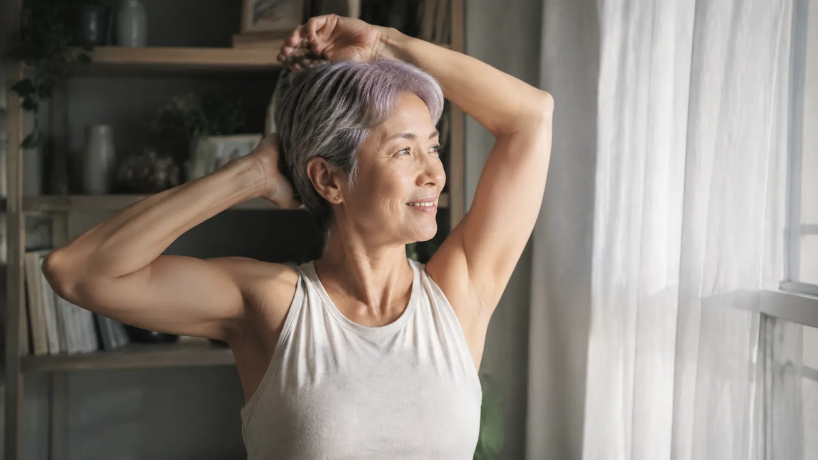 Woman in her 60s stretching gently in morning sunlight by a window, looking rested