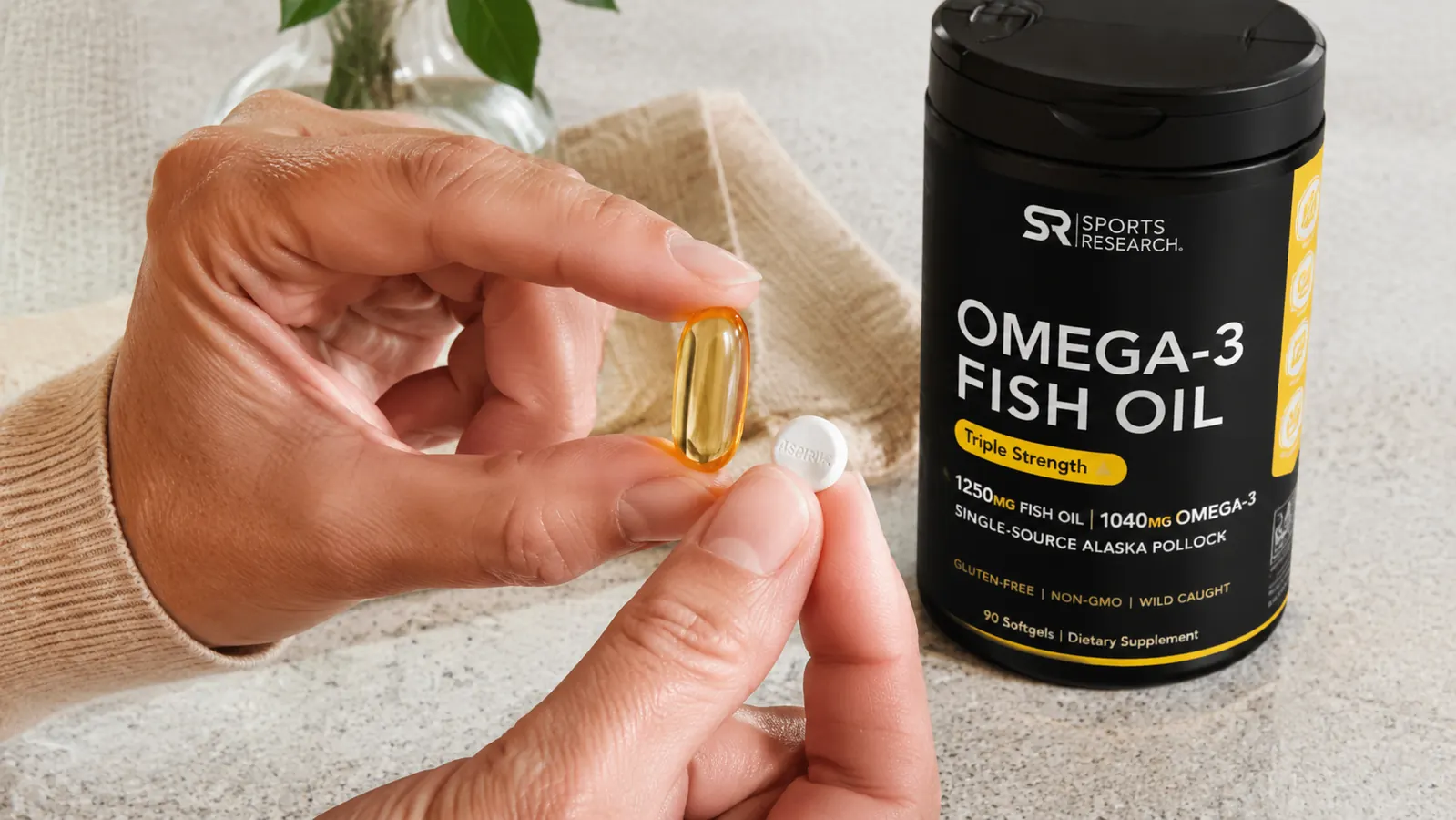 Close-up of a hand holding a large amber softgel capsule next to a standard aspirin tablet to show size comparison