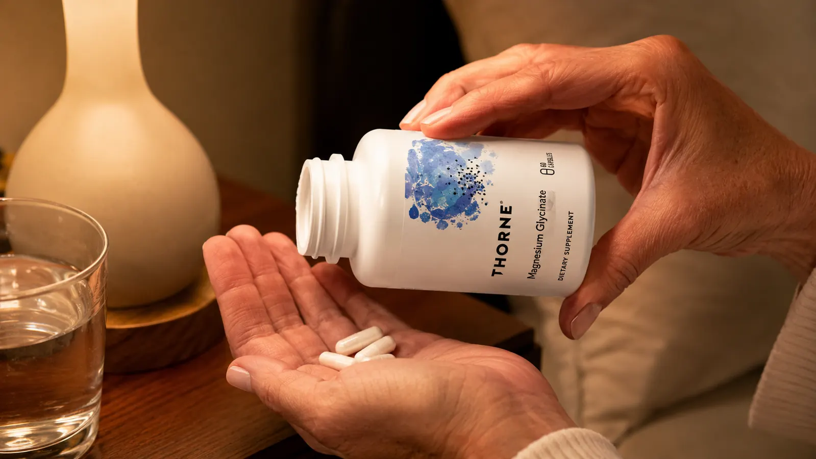 A woman's hand tipping two white capsules from a supplement bottle into her palm above a nightstand